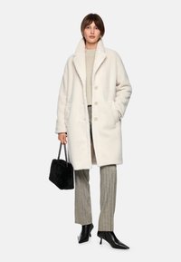 Weißer Kunstpelzmantel mit großen Knöpfen, Seitentaschen und Schalkragen. Kombiniert mit grauen gestreiften Hosen und schwarzen spitzen Absatzschuhen.