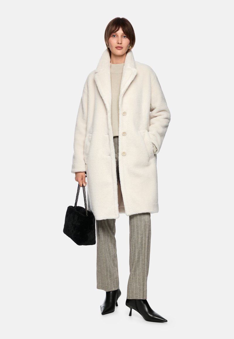 Weißer Kunstpelzmantel mit großen Knöpfen, Seitentaschen und Schalkragen. Kombiniert mit grauen gestreiften Hosen und schwarzen spitzen Absatzschuhen.