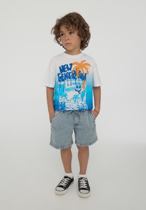 T-shirt bianca a maniche corte con grafica di palme blu e arancioni, abbinata a pantaloni corti di jeans e sneakers nere con suole e lacci bianchi.