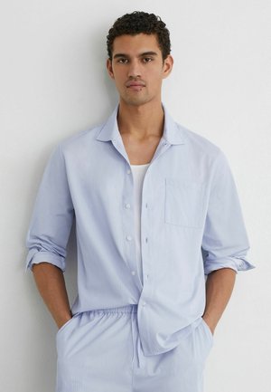 Fiatal férfi áll fehér fal előtt, világoskék csíkos gombos inggel, fehér felső alatt, hozzáillő, húzózsinóros nadrágban.