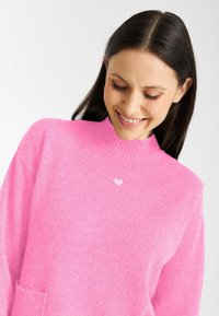 Różowy sweter z wysokim dekoltem, żebrowanym wykończeniem i teksturowanym materiałem. Posiada mały biały detal w kształcie serca na piersi.