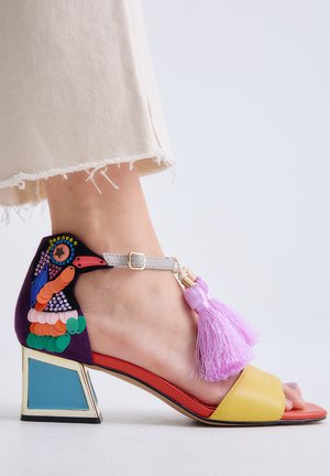 Sandalia de punta abierta en el pie con un tacón bajo de bloque, decoración colorida de pájaro en el talón y borlas rosas en la correa del tobillo.
