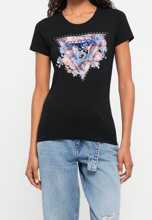 Kobieta w czarnym dopasowanym T-shircie z ćwiekowanym logo Guess w kształcie trójkąta otoczonym fioletowymi i różowymi kwiatami, w zestawieniu z jasnoniebieskimi dżinsami.