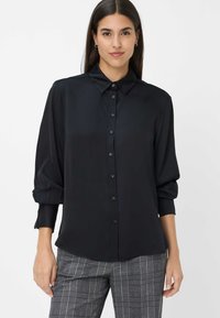 Camicia nera a maniche lunghe con bottone, realizzata in tessuto morbido con colletto strutturato, dotata di bottoni sul fronte e polsini arricciati.