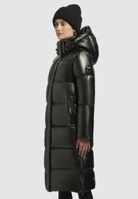 Svart vadderad lång kappa med en glänsande yta, stor huva, dragkedjefickor på sidorna och en logotyp-patch på ärmen. Mjuk textur.