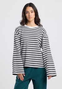 Jonge vrouw met lang, donker haar, gekleed in een losse zwart-witte gestreepte langemouwen top en teal kleurige broek, staande tegen een effen achtergrond.