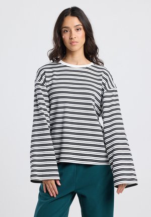 Jonge vrouw met lang, donker haar, gekleed in een losse zwart-witte gestreepte langemouwen top en teal kleurige broek, staande tegen een effen achtergrond.