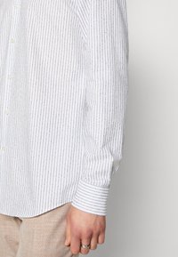 Man in een witte blouse met lange mouwen met dunne marineblauwe verticale strepen en beige broek, met een arm en hand in beeld waarop een zilveren ring om de ringvinger te zien is.