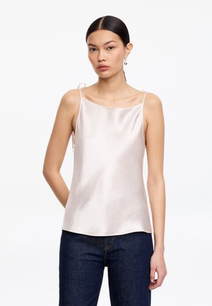 Kvinde med mørkt hår samlet i en lav hestehale, iført perleøreringe og en skinnende blid pink satin camistop kombineret med mørkeblå jeans.