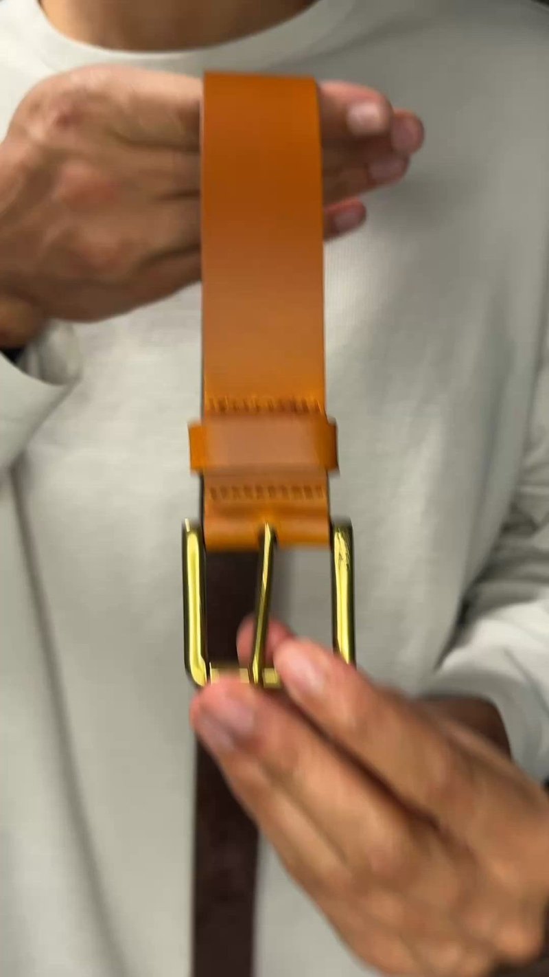 Manos sosteniendo un cinturón de cuero color canela con una hebilla dorada, colocado verticalmente frente a una persona que lleva una camisa de color claro.