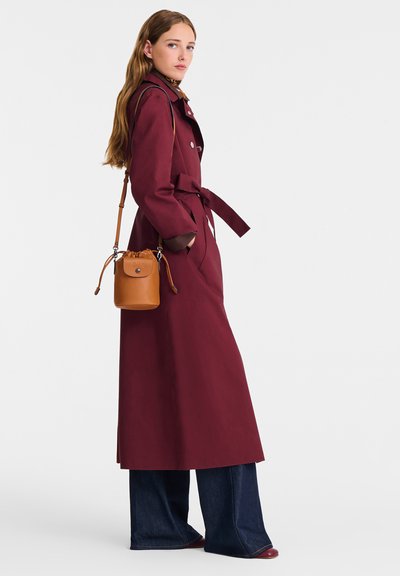 Burgundy trench coat with a belted waist, deep collar, and long sleeves, paired with a small brown leather crossbody bag and wide-leg jeans.