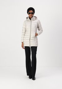Giubbotto piumino grigio chiaro con cuciture orizzontali, zip frontale, cappuccio e tasche laterali, indossato sopra pantaloni neri e stivali.
