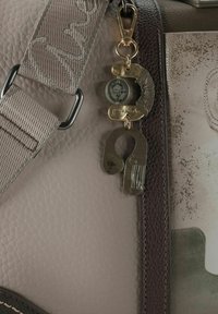 Graue, strukturierte Tasche mit glatter Oberfläche, verziert mit einem dekorativen Schlüsselanhänger mit metallischen Akzenten und einem runden Anhänger. Verstellbarer Riemen inklusive.