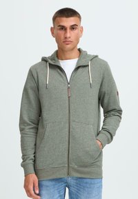 Zeleni zip-up dukserica izrađena od mekanog materijala, s kapuljačom na vezanje, prednjim džepovima i suptilnom heathered teksturom.