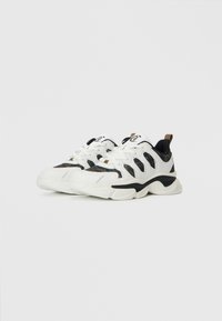 Weiße und schwarze klobige Sneakers mit Camouflage-Mustern und beigen Zuglaschen, nebeneinander auf einem weißen Hintergrund präsentiert.