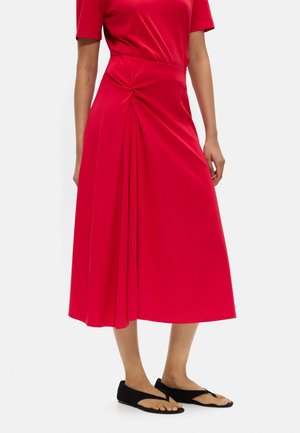 Kobieta nosząca czerwoną spódnicę midi z marszczonym węzłowym detalem po lewej stronie, w połączeniu z czarnymi sandałami z odkrytymi palcami.
