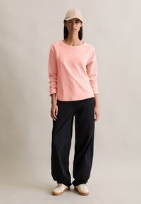 Helles rosa Langarmhemd mit rundem Ausschnitt, kombiniert mit lockeren schwarzen Hosen und beigen Sneakern. Eine beigefarbene Mütze rundet das Outfit ab.