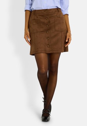 Mini-jupe en daim marron avec fermeture à boutons et deux poches plaquées, présentant une coupe droite et une texture douce. Associée à des chaussures noires.