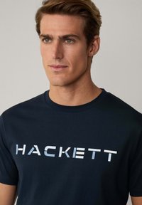 Dunkelblaue Baumwollt-Shirt mit dem Wort "HACKETT" in einem Farbverlauf von Weiß zu Blau. Klassischer Rundhalsausschnitt, kurze Ärmel, glatte Textur.