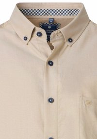 Beige Hemd mit Knöpfen aus strukturiertem Stoff. Blaue Knöpfe und ein kariertes Muster im Inneren des Kragens. Kleinere Brusttasche.