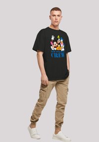 Juoda trumpa rankovėmis marškinėliai su ryškiu Disney personažo grafiku ir tekstu "Disney CREW"; derinami su smėlio spalvos cargo kelnėmis ir baltomis sportinėmis bateliais.