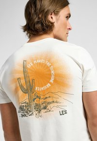 Camiseta blanca con un diseño ilustrado de un cactus y un personaje, con un fondo de degradado naranja y el texto "LEE HACE QUE EL SOL BRILLE MÁS".