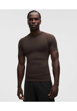 Uomo atletico con capelli intrecciati che indossa una maglietta aderente a maniche corte marrone scuro e pantaloncini neri su uno sfondo chiaro uniforme.