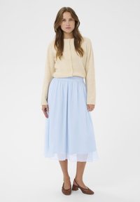 Světle modrá sukně po kolena s volným střihem a elastickým pasem, kombinovaná s béžovým svetrem na knoflíky vpředu a hnědými pletenými podpatky.