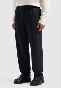 Marineblauwe broek met plooien, slim fit, elastische tailleband en een gladde stofstructuur. Gecombineerd met zwarte veterschoenen.