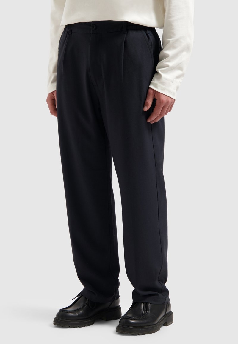 Marineblauwe broek met plooien, slim fit, elastische tailleband en een gladde stofstructuur. Gecombineerd met zwarte veterschoenen.