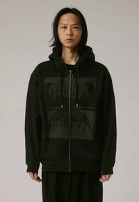 Jovem com cabelo longo e escuro veste um hoodie preto com fecho que apresenta um design abstrato subtil na parte da frente, contra um fundo simples.