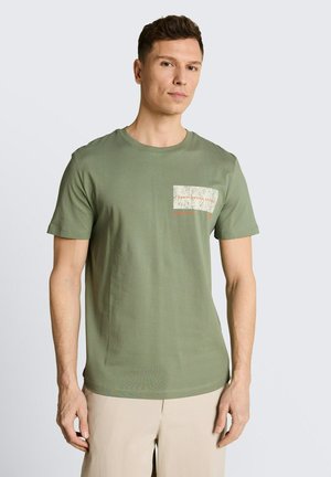 Mężczyzna stojący, ubrany w oliwkową koszulkę z krótkim rękawem z kieszonką na piersi oraz jasnobeżowe spodnie, neutralne tło.