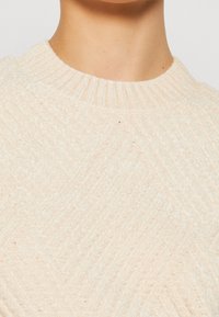 Gros plan sur une personne portant un pull en tricot texturé de couleur crème avec un col rond côtelé.
