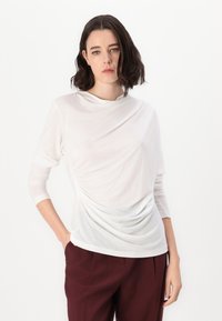 White long-sleeve top with gathered texture on one side, crew neckline, and soft fabric. Paired with burgundy wide-leg trousers.