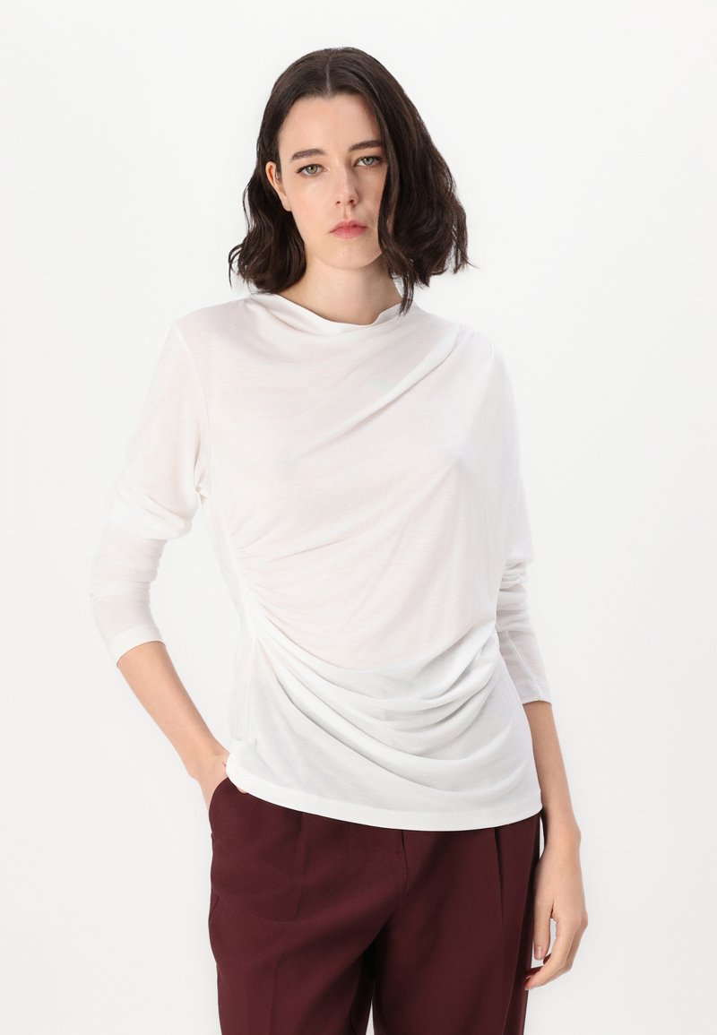 Biała bluzka z długim rękawem o marszczonym wykończeniu z jednej strony, z okrągłym dekoltem i miękkiej tkaniny. Połączona z bordowymi spodniami typu wide-leg.