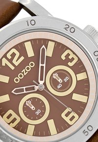 Montre ronde avec un cadran marron. Index et aiguilles dorés. Deux sous-cadrans. Boîtier argenté. Bracelet en cuir marron avec des détails de couture.