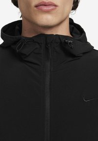 Zwarte Nike-jas met een rits aan de voorkant, verstelbare capuchon met trekkoorden, en een subtiel logo op de linkerborst. Gladde textuur.