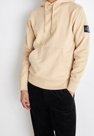 Person trägt beige Kapuzenpullover mit Fronttasche und Logo-Patch am Ärmel, kombiniert mit schwarzen strukturierten Hosen.