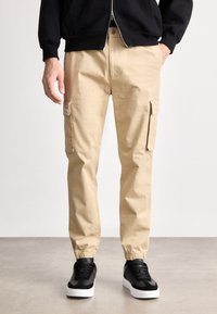 Beige cargobroek met zijzakken, elastische enkelmanchetten en een duurzaam materiaal. Gecombineerd met zwarte sneakers met witte zolen.
