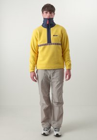 Geel fleece sweatshirt met een hoge kraag, marineblauwe rits en een decoratieve bloemband, gecombineerd met lichtgrijze broek en zwarte sportschoenen.