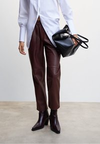 Personne portant un pantalon en cuir marron foncé, des bottines à talons assorties, une chemise blanche à manches longues, tenant un grand sac à main noir.