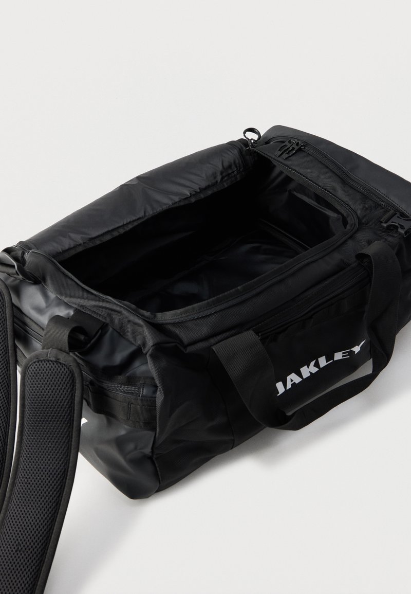 Sort Oakley duffel taske med åben hovedrum, dobbelte håndtag, skulderrem og synligt hvidt logo på sidelommen.