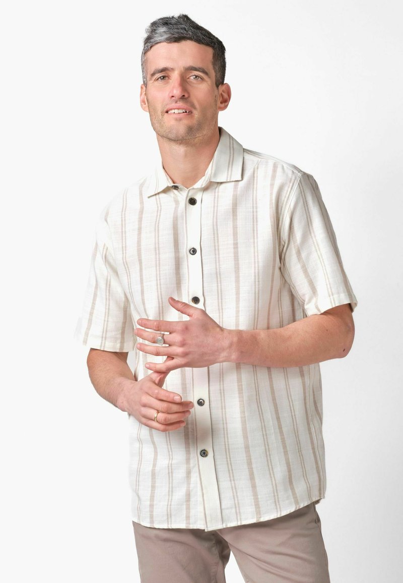 Homme portant une chemise à rayures beige à manches courtes et un pantalon de couleur claire, ajustant sa bague sur un fond blanc.