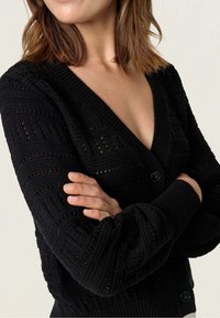 Žena s hnědými vlasy po ramena, oblečená v černém pleteném svetru s texturou, s hlubokým výstřihem do V a knoflíky, s překříženýma rukama.