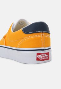 Keltainen kangaskenkä sinisillä nahkaosilla, valkoisella välipohjalla ja punaisella, jossa lukee "VANS OFF THE WALL." Kontrastiosoitus näkyvissä.