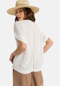 Top in tessuto bianco stropicciato con maniche corte, caratterizzato da un bottone sul retro e una vestibilità rilassata, abbinato a pantaloni corti marroni e un cappello di paglia.