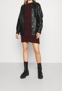 Veste en cuir noir boutonnée portée sur une robe en maille côtelée bordeaux, associée à des bottes noires montantes à semelle épaisse.