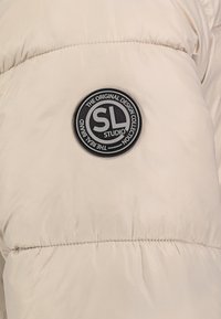 Beige gewatteerde jas met een gladde textuur. Heeft een ronde zwarte logopatch met de tekst "SL STUDIO" in het wit.