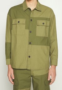 Camicia verde oliva con bottoni in cotone, dotata di due tasche sul petto, colletto e toppe a contrasto in diverse tonalità di verde.