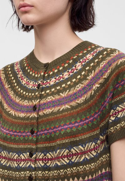 Femme portant un pull en maille vert boutonné avec des motifs géométriques et floraux multicolores, manches courtes et encolure ronde.